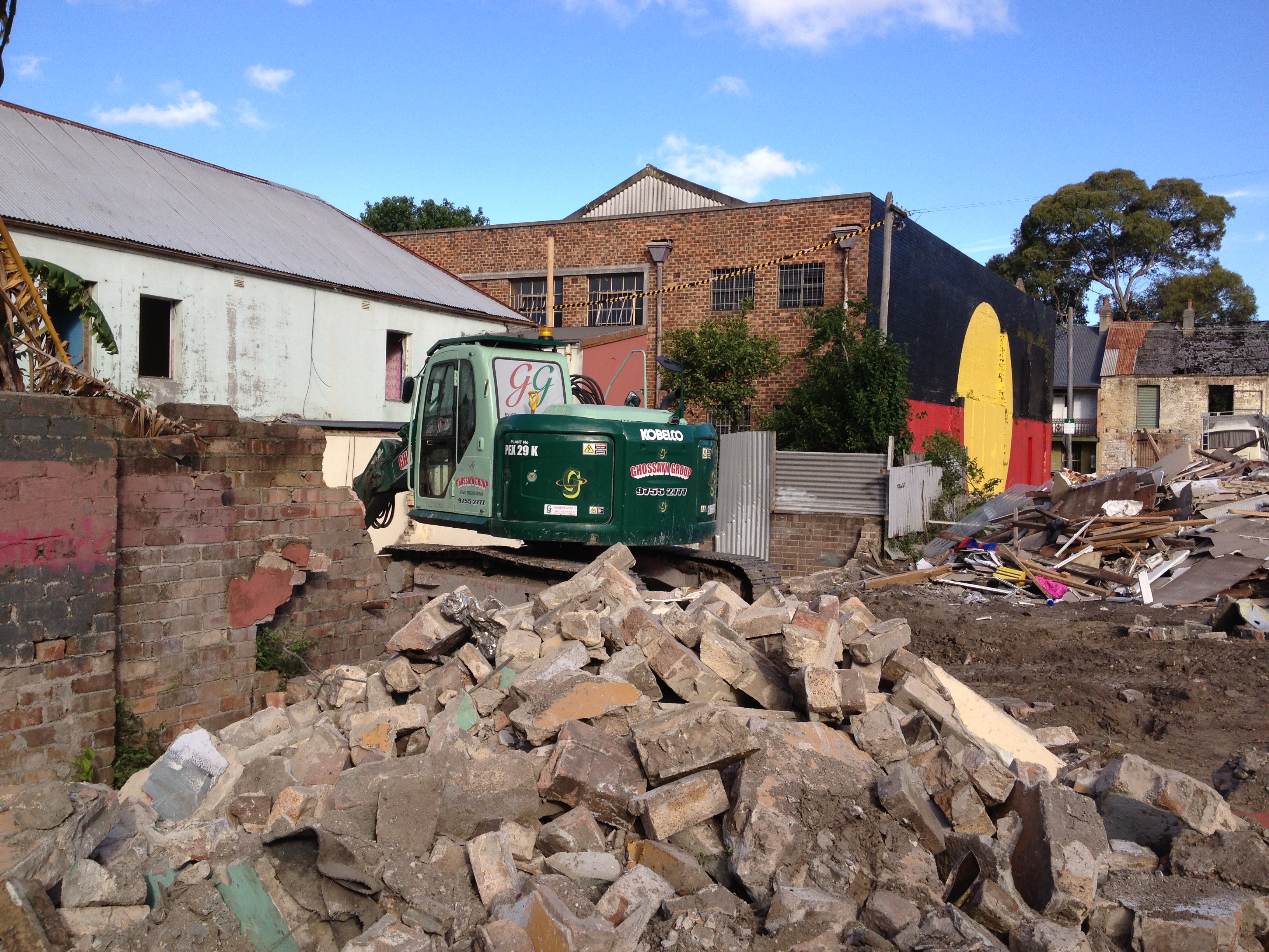Foreground of the image is a pile of bricks and a demolition machine and in the background can see a building with the Aboriginal Flag painted on the side