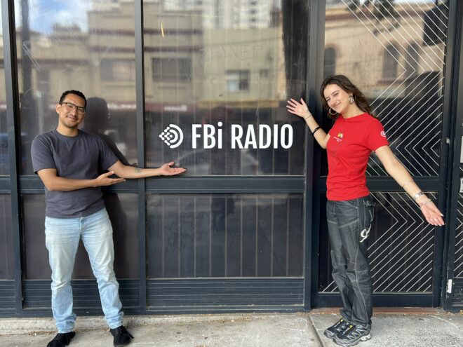 Outgoing host Kwame and new host Gabriella standing in front of the fbi radio door.